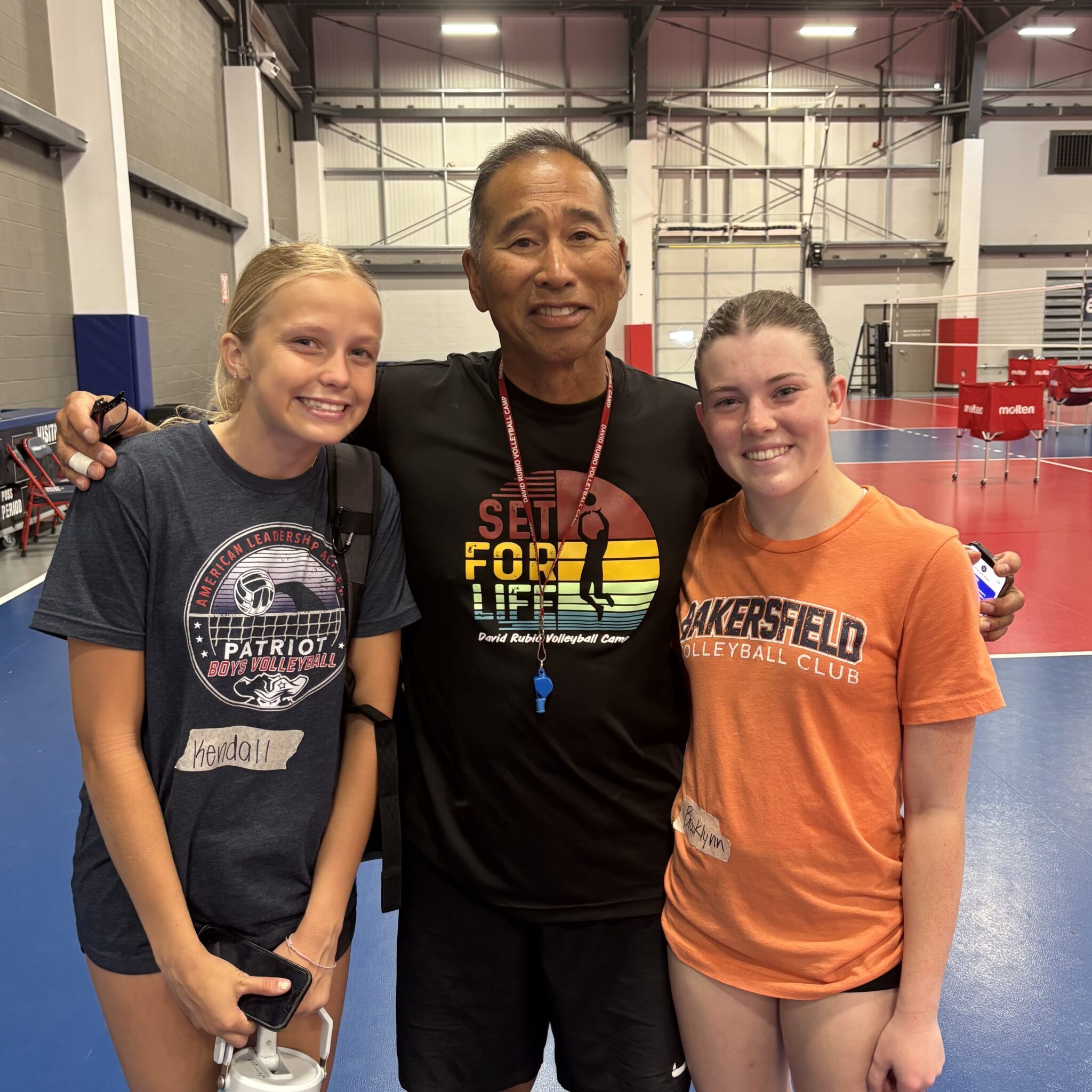 The image shows a man standing between two young women, all smiling and posing for a photo. They are likely at a volleyball camp, as indicated by their shirts and the surrounding court.